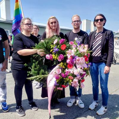 Vier Menschen stehen im Freien mit einem großen Kranz aus bunten Blumen, darunter Helge Langer, hinter ihnen eine Regenbogenfahne