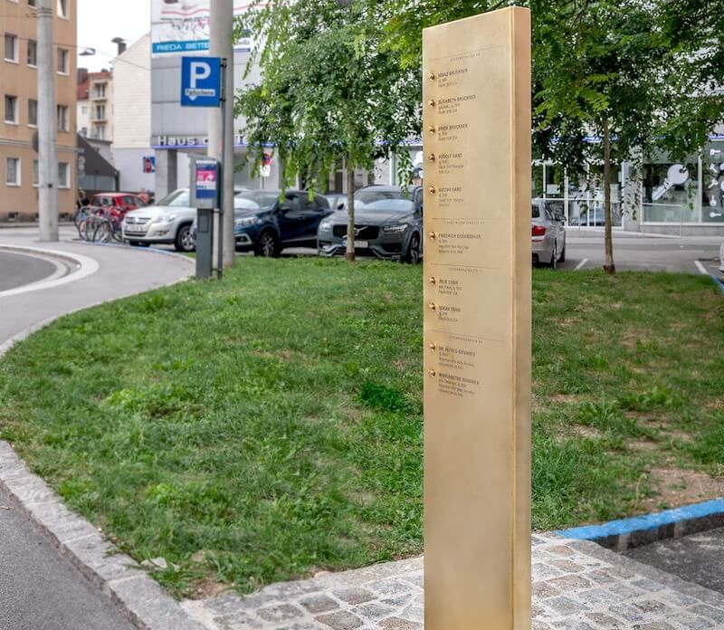 Eine Erinnerungsstele aus Messing mit eingravierten Namen steht unter Bäumen an einer Linzer Straßenecke