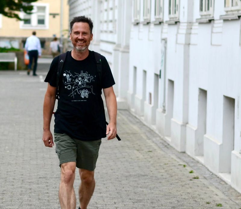 Helge Langer geht lächelnd auf einem breiten, gepflasterten Gehsteig an einer hellen Hausfassade entlang