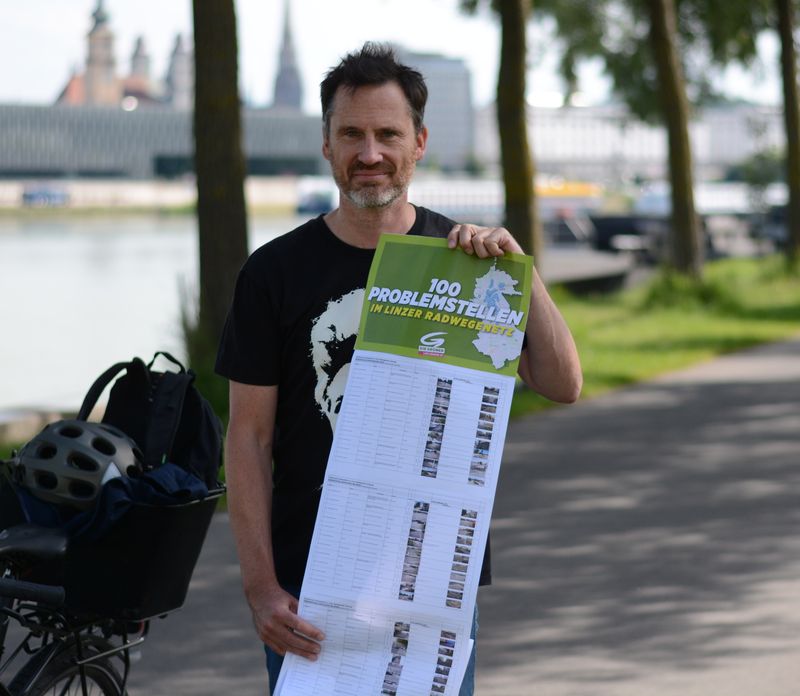 Helge Langer an der Donau mit dem langen Plakat „100 Problemstellen im Linzer Radwegenetz“, daneben sein Fahrrad mit Helm im Korb