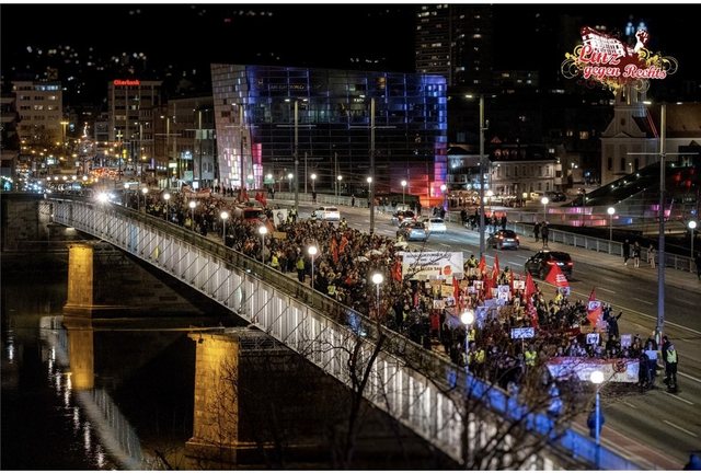 Ein langer Demonstrationszug mit Fahnen und Transparenten zieht bei Nacht über eine beleuchtete Donaubrücke, dahinter das Ars Electronica Center