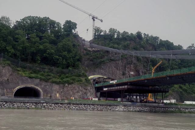 Blick über die Donau auf die Urfahrwänd, links ein Tunnelportal im Fels, rechts das Stahlgerüst der Brückenbaustelle mit Seilen und einem Baukran auf dem Hang