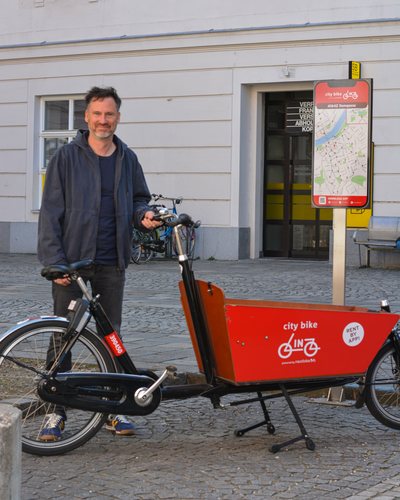 Helge Langer steht lächelnd hinter einem roten Leih-Lastenrad von city bike Linz, daneben eine Stationstafel mit Stadtplan vor einer hellen Hausfassade