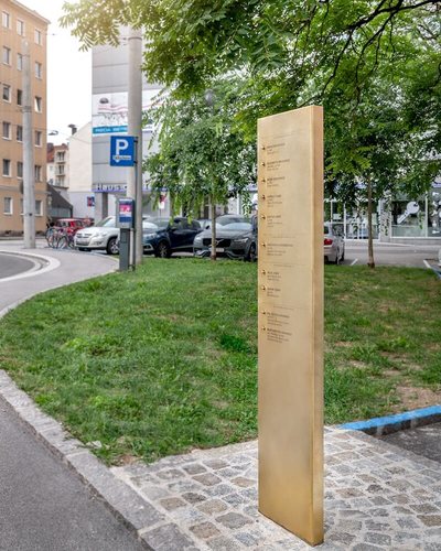 Eine schmale Stele aus Messing auf einer Rasenfläche, darauf untereinander eingravierte Namen, neben jedem Namen ein kleiner Klingelknopf