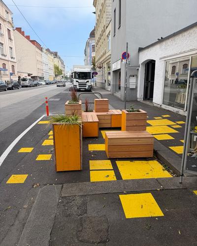 Hölzerne Pflanztröge und Sitzbänke stehen auf einer gelb markierten Fläche am Fahrbahnrand einer Straße mit Geschäften