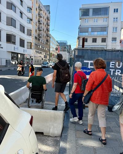 Christoph Etzlstorfer, Vizepräsident des Österreichischen Behindertensportverbands, fährt im Rollstuhl auf eine schmale Lücke zwischen einer Baustellenabsperrung aus Beton und einem parkenden Auto zu, neben ihm weitere Teilnehmende des Rundgangs