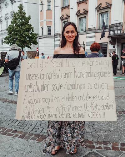 Eine Frau hält auf dem Hauptplatz ein handgeschriebenes Schild: „Soll die Stadt Linz die Verbauung unseres Grüngürtels/unserer Naherholungsgebiete unterbinden sowie Zuzahlungen zu allen Autobahnprojekten einstellen und dieses Geld stattdessen für öffentlichen Verkehr einsetzen?“ und darunter „Mehr als 7000 Linzer wollen gefragt werden!!!“