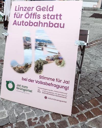 Ein großes Plakat auf Kopfsteinpflaster mit der Aufschrift „Linzer Geld für Öffis statt Autobahnbau“ und „Stimme für Ja! bei der Volksbefragung“ des Bündnisses „Ja! zum Grüngürtel“