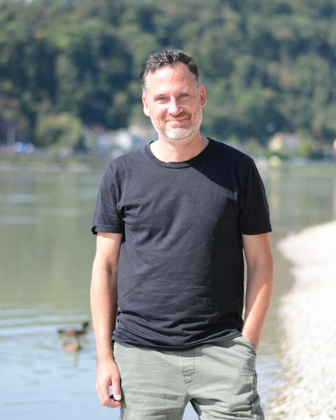 Helge Langer steht lächelnd am Donaustrand, hinter ihm das Wasser und bewaldete Hügel