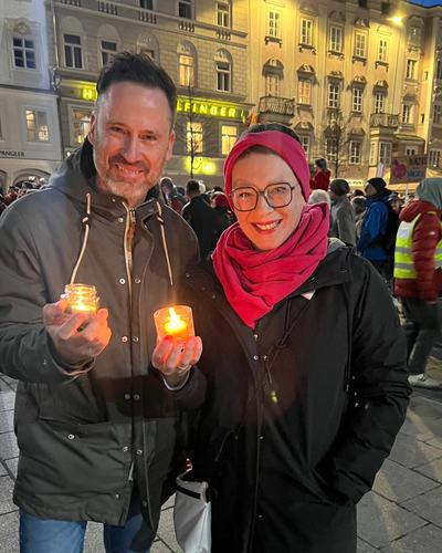 Helge Langer und eine Frau mit rotem Kopftuch halten am Abend auf dem Linzer Hauptplatz voller Menschen je ein Glas mit einer brennenden Kerze