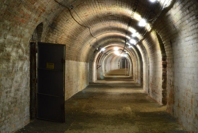 Ein langer, gemauerter Gang mit gewölbter Ziegeldecke, beleuchtet von Lampen an der Decke, links eine offene Metalltür