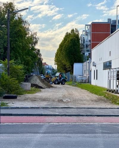 Eine geschotterte Trasse zwischen Bäumen und einem Gewerbebau, darauf ein Erdhaufen und ein kleiner Radlader, vorne eine Straße mit rotem Radweg