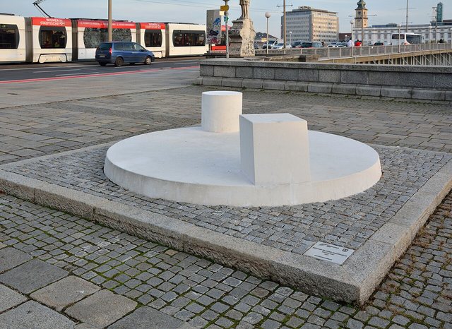Eine flache, runde weiße Steinscheibe auf einem gepflasterten Platz, darauf ein weißer Zylinder und ein weißer Würfel, im Hintergrund eine Straßenbahn und eine Brücke