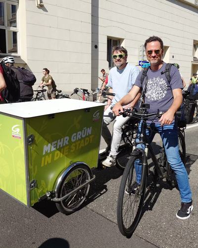 Helge Langer mit Sonnenbrille auf seinem Fahrrad zwischen weiteren Radfahrenden auf einer Straße, daneben ein grünes Lastenrad mit der Aufschrift „Mehr Grün in die Stadt“