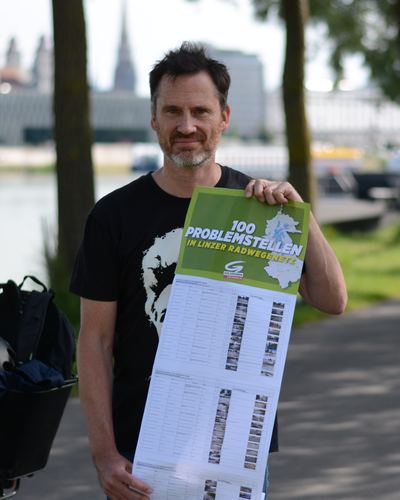 Helge Langer hält ein langes Plakat mit der Überschrift „100 Problemstellen im Linzer Radwegenetz“ und einer Tabelle mit Fotos der einzelnen Stellen, hinter ihm die Donau