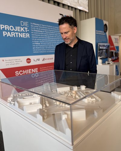 Helge Langer betrachtet in einer Ausstellung zur Regional-Stadtbahn ein weißes Architekturmodell unter einer Glashaube, hinter ihm eine Tafel über die Projektpartner