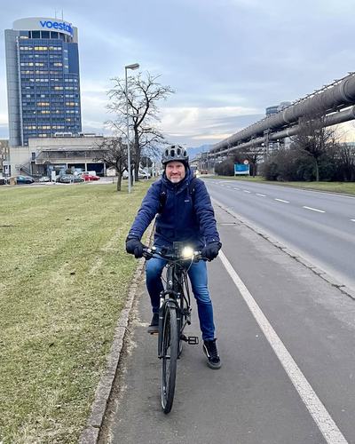 Helge Langer fährt mit Helm und Licht auf einem Radweg neben einer breiten Straße, im Hintergrund das Hochhaus der voestalpine und eine Rohrleitungsbrücke
