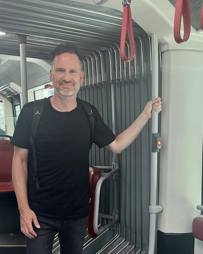 Helge Langer steht lächelnd in einer Linzer Straßenbahn und hält sich an einer Haltestange fest