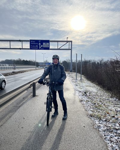 Helge Langer steht mit Helm und Rad auf dem Geh- und Radweg neben der Fahrbahn, darüber ein Wegweiser zur A7 nach Wien und Salzburg, am Rand Raureif