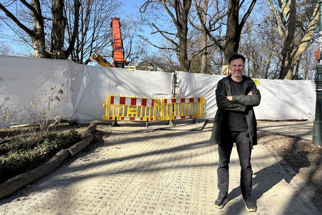 Helge Langer steht mit verschränkten Armen vor einem weißen Bauzaun mit rot-gelben Absperrungen, dahinter kahle Bäume und ein Kran